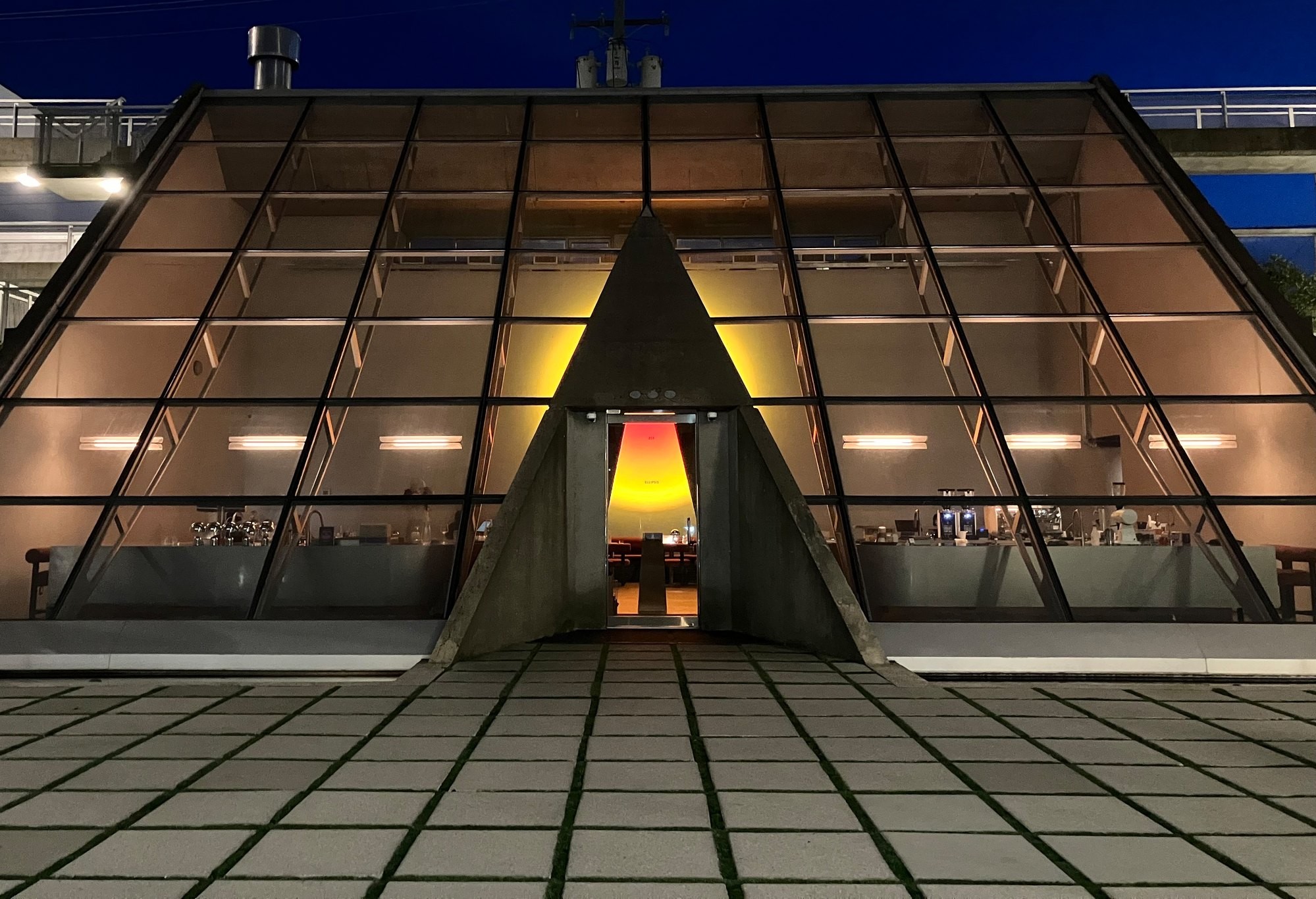 A glass A-frame pyramid building at blue hour, its triangular entrance glowing amber from within